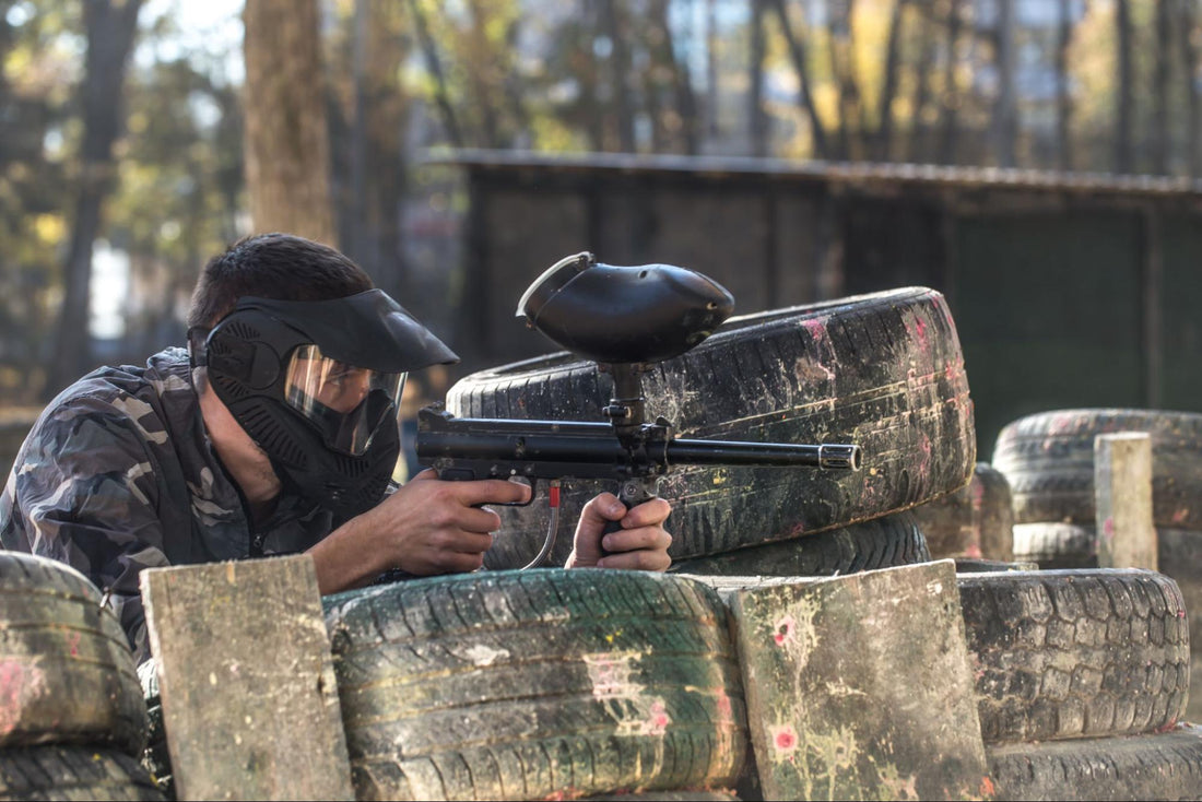 Player in protective gear aiming airsoft shotgun behind tire barricade during gameplay at Canadian airsoft field